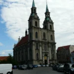 Barokowy kościół jezuitów w Brzegu z dwiema wysokimi wieżami i jasną elewacją, parking przed świątynią w słoneczny dzień