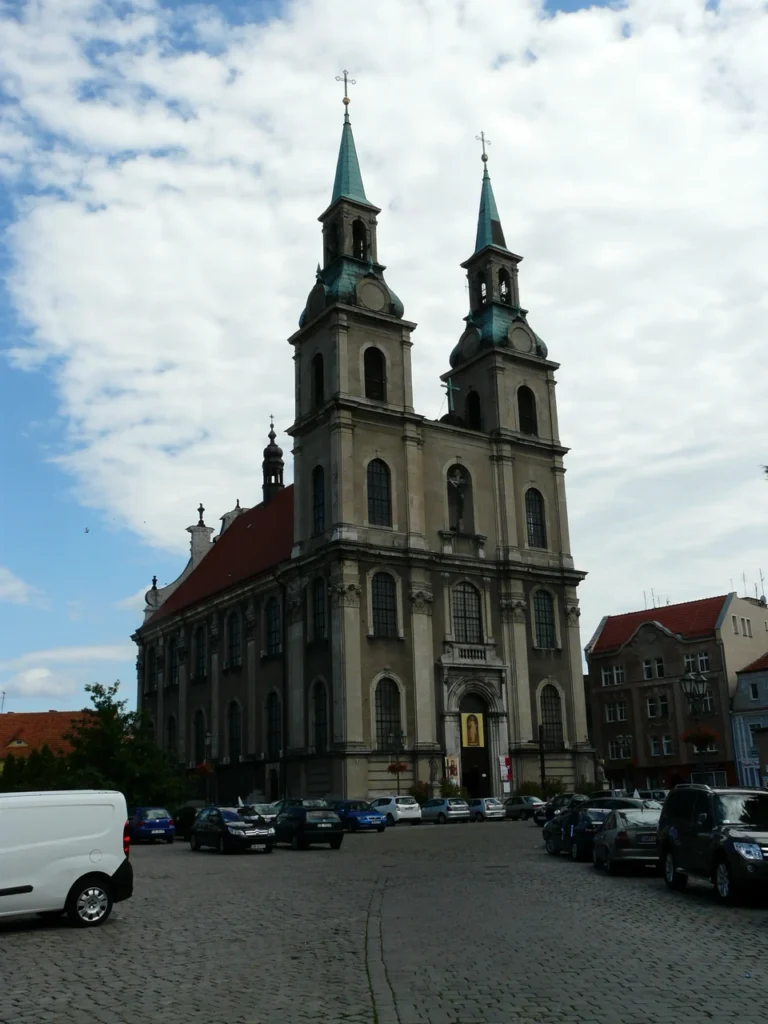 Barokowy kościół jezuitów w Brzegu z dwiema wysokimi wieżami i jasną elewacją, parking przed świątynią w słoneczny dzień