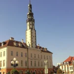 Ratusz w Grodkowie z wysoką wieżą zegarową, stojący pośrodku rynku, otoczony kamienicami