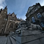 Kamienna rzeźba lwa na schodach przed zamkiem w Mosznej, z widokiem na fasadę pałacu w szerokim ujęciu