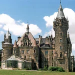 Zamek w Mosznej widziany z boku, rozbudowana bryła z wieżami na tle nieba i trawnikiem na pierwszym planie