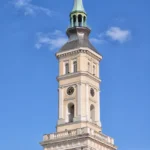 Wieża ratusza w Prudniku zakończona iglicą, z czterema tarczami zegara widocznymi nad dachem budynku