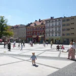Rynek w Brzegu z fontannami wodnymi i spacerującymi ludźmi, w tle zabytkowe kamienice i słoneczny letni dzień
