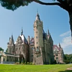 Zamek w Mosznej widziany z parku, z wysoką kamienną wieżą i rozłożystym drzewem na pierwszym planie