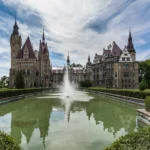 Zamek w Mosznej widziany zza fontanny w parku, z rozświetloną fasadą i wodą na pierwszym planie