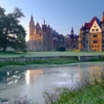 Zamek w Mosznej oświetlony porannym słońcem, odbijający się w stawie parkowym na pierwszym planie